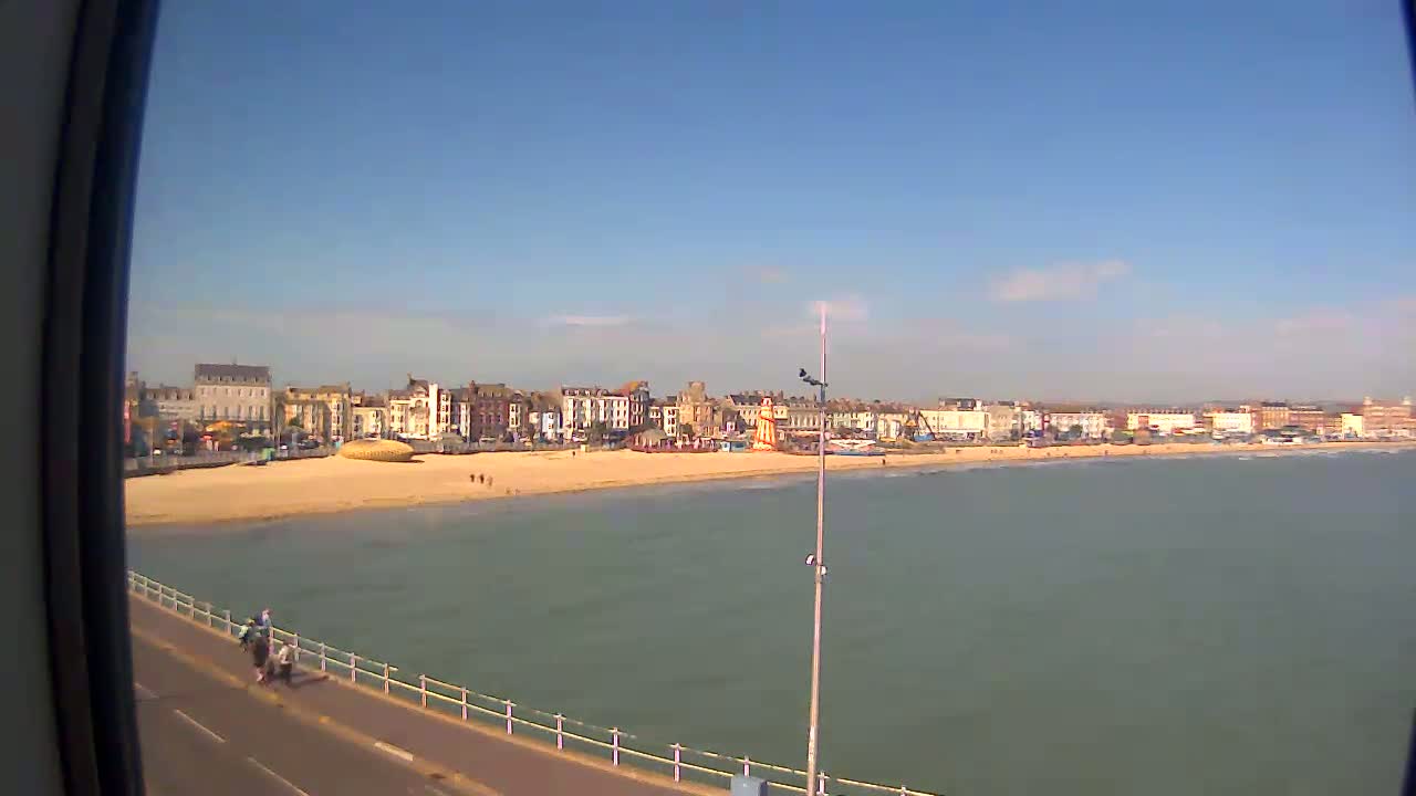 A sandy beach with buildings and a pier stretches along a calm, light-green ocean under a clear blue sky.