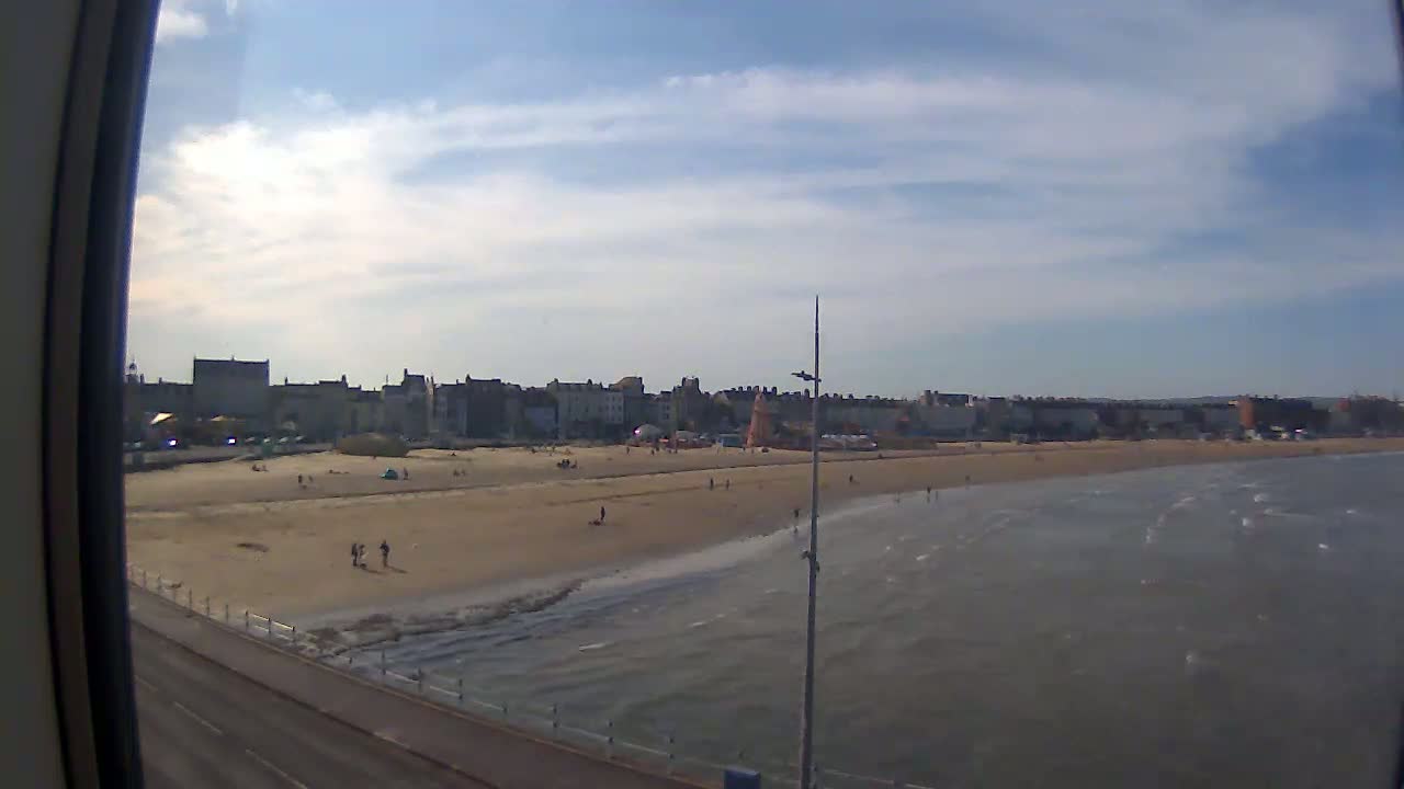 A sandy beach with a few people on it stretches in front of a town under a partly cloudy sky, with calm, slightly rippled water lapping the shore.