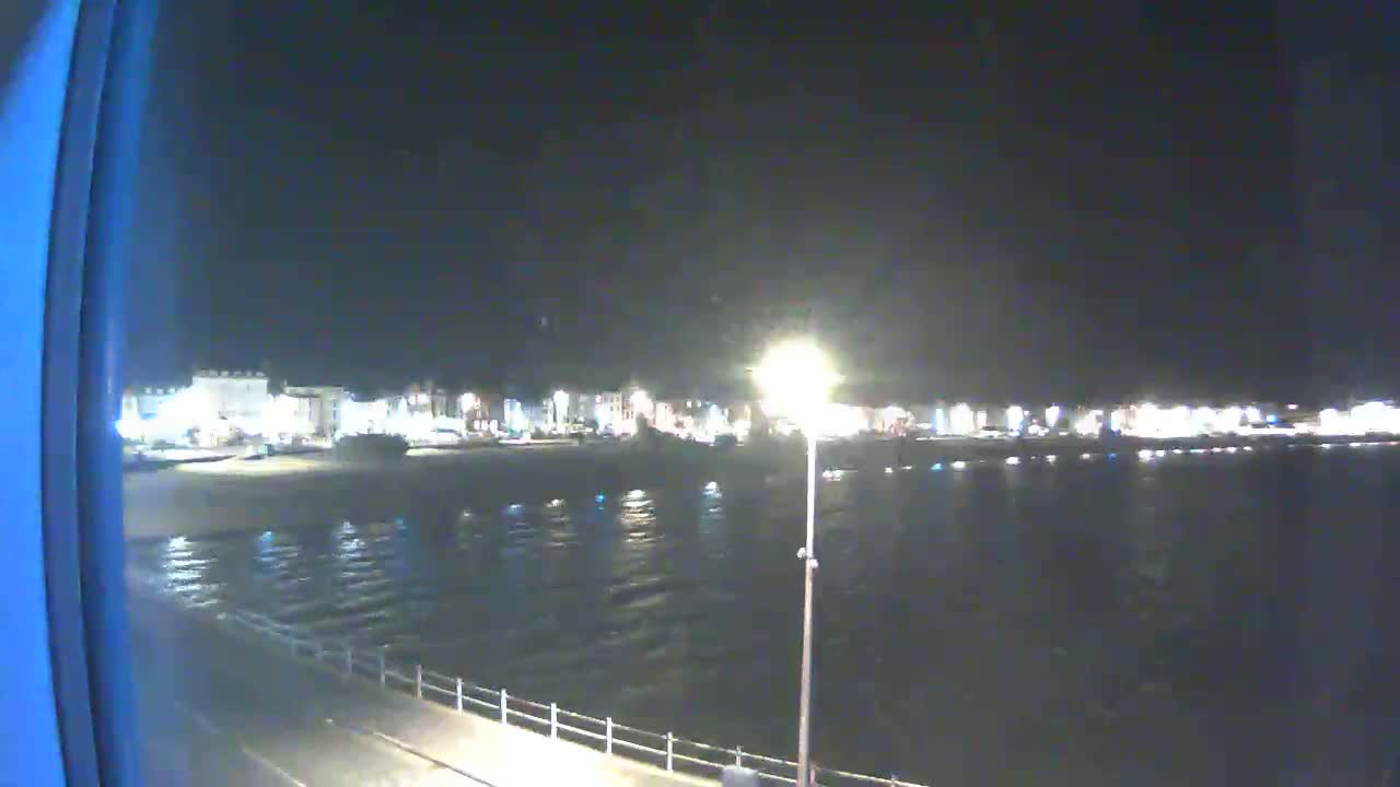A nighttime view of a calm body of water reflecting the lights of a town across the shore, with a seawall and street visible in the foreground.