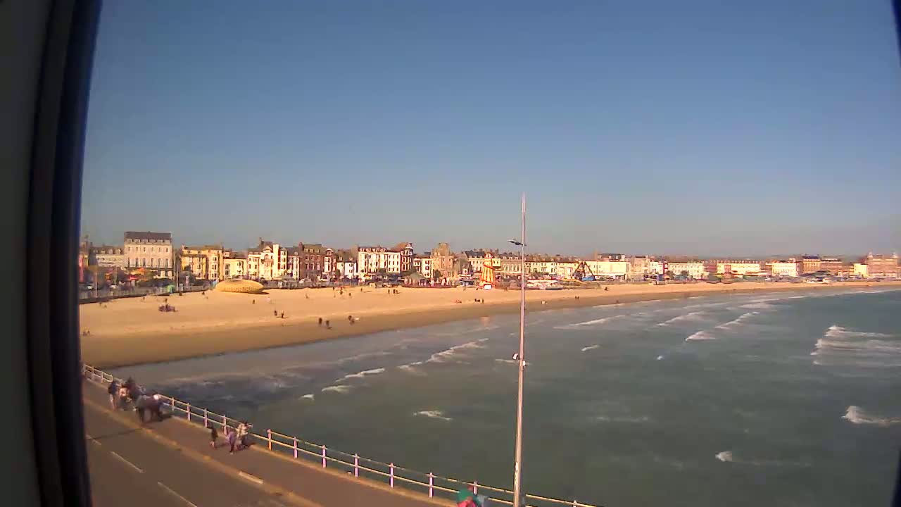 A sandy beach with a row of buildings and a Ferris wheel behind it is viewed from a pier on a sunny day with moderate waves in the ocean.