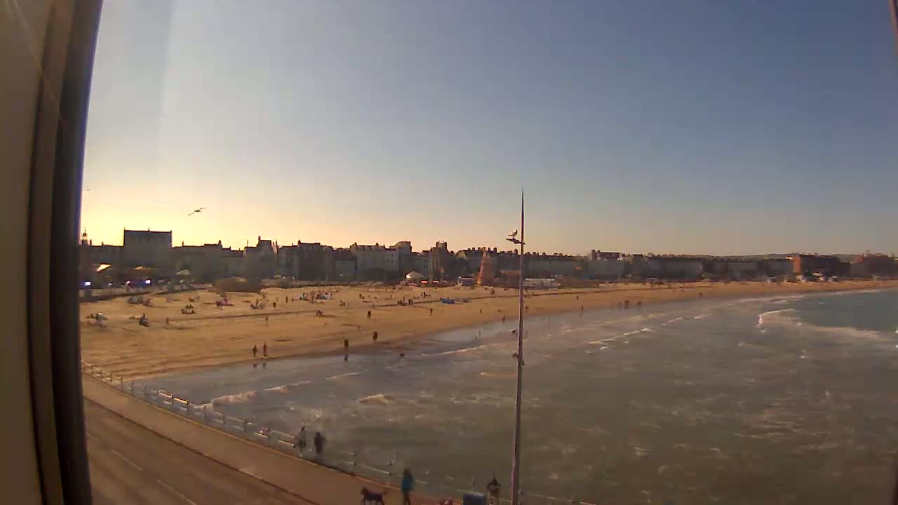 A sandy beach with scattered people, gentle waves, and a town of buildings in the background under a clear, sunset sky.