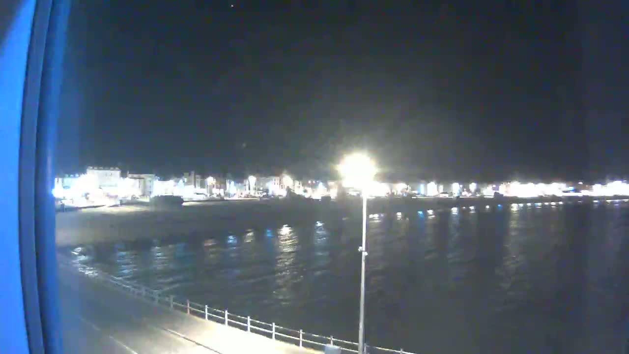 A nighttime view of a calm body of water next to a dimly lit beachfront town under a clear, dark sky.