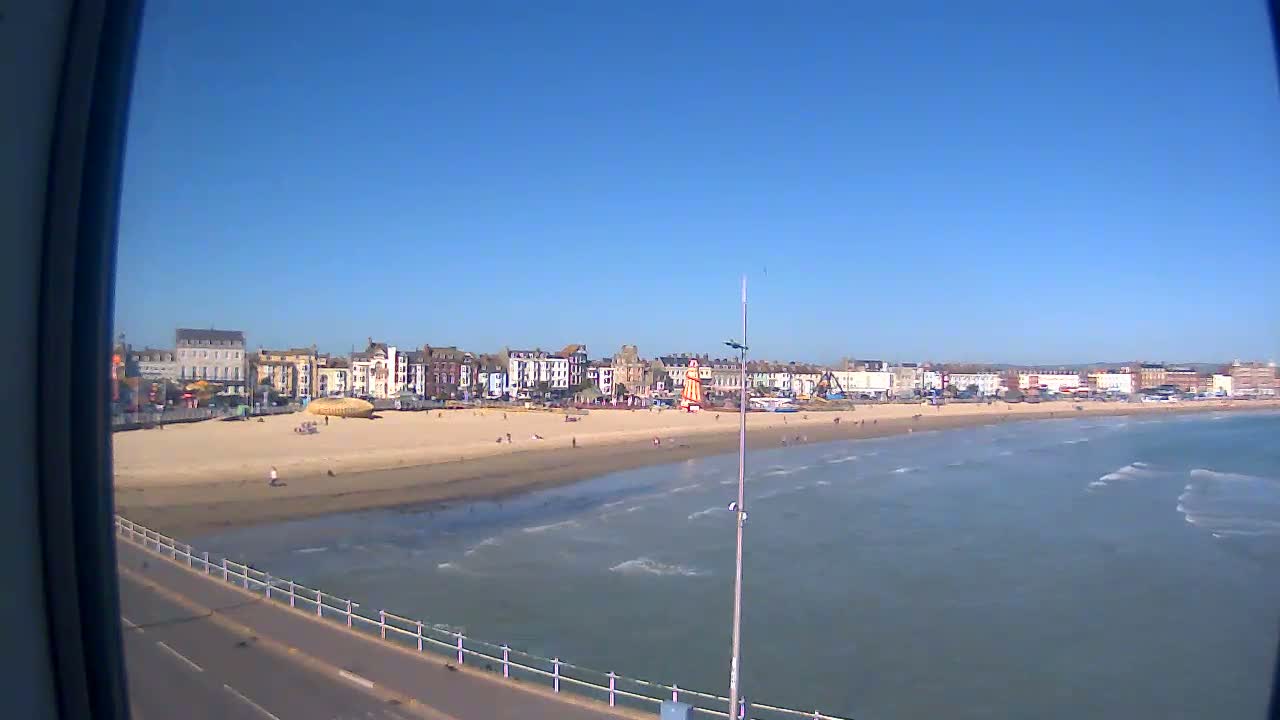 A sandy beach with a row of buildings behind it stretches along a calm, blue ocean under a clear, sunny sky.