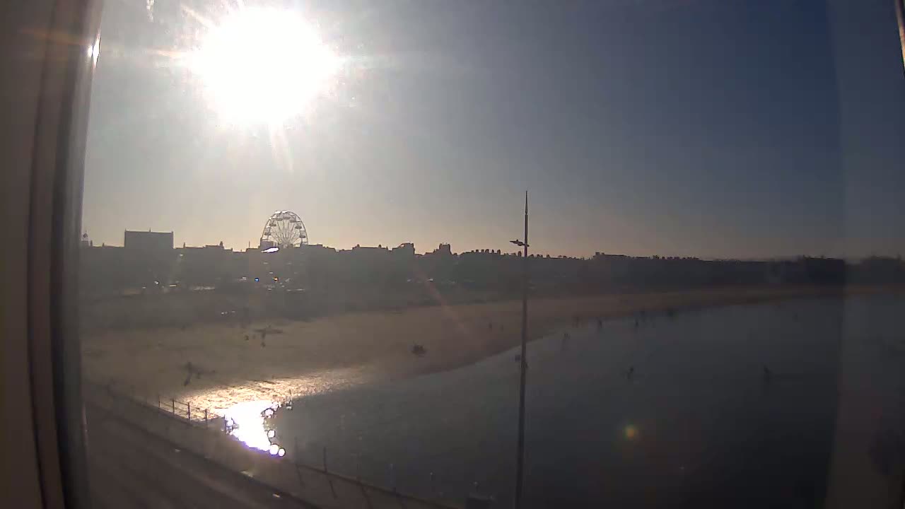 A sunny day at a beach shows a Ferris wheel and buildings in the background, with calm water lapping a sandy shore where a few people are visible.