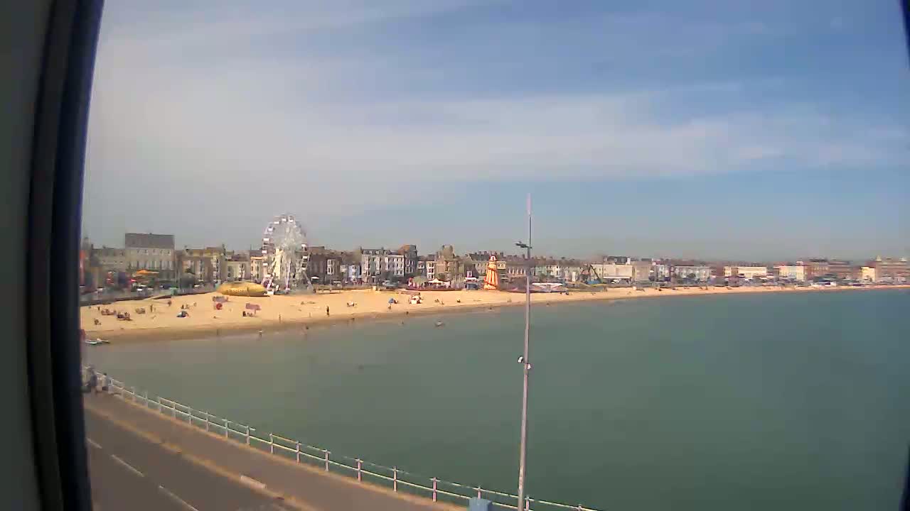 A sandy beach with a Ferris wheel and buildings stretches along a calm, greenish-blue sea under a mostly clear sky.