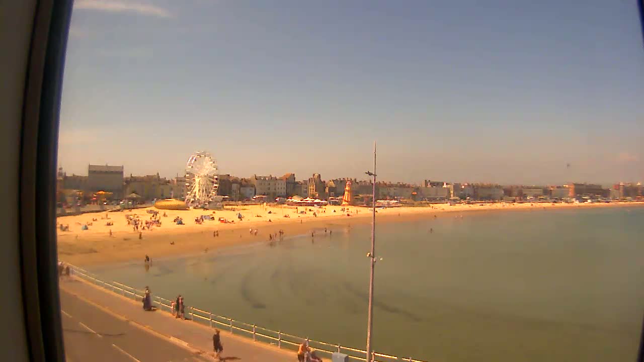 A sunny day at a beach shows a Ferris wheel and other fairground rides, a sandy beach with people, and calm, shallow water.