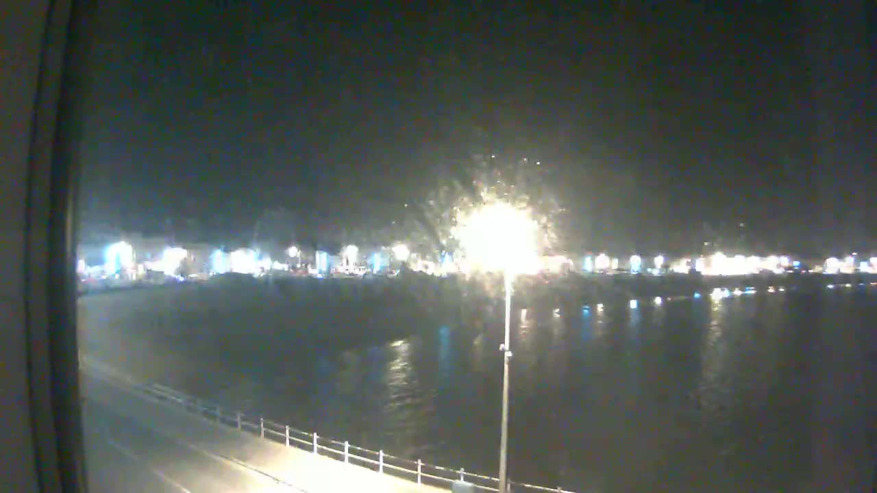 A nighttime view of a calm body of water reflecting the lights of a town across a paved waterfront.