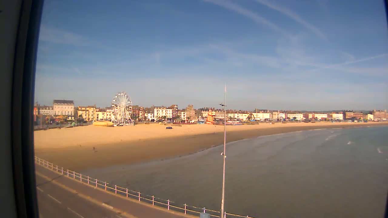 A sandy beach with a Ferris wheel and buildings behind it stretches along a calm ocean under a mostly clear blue sky.