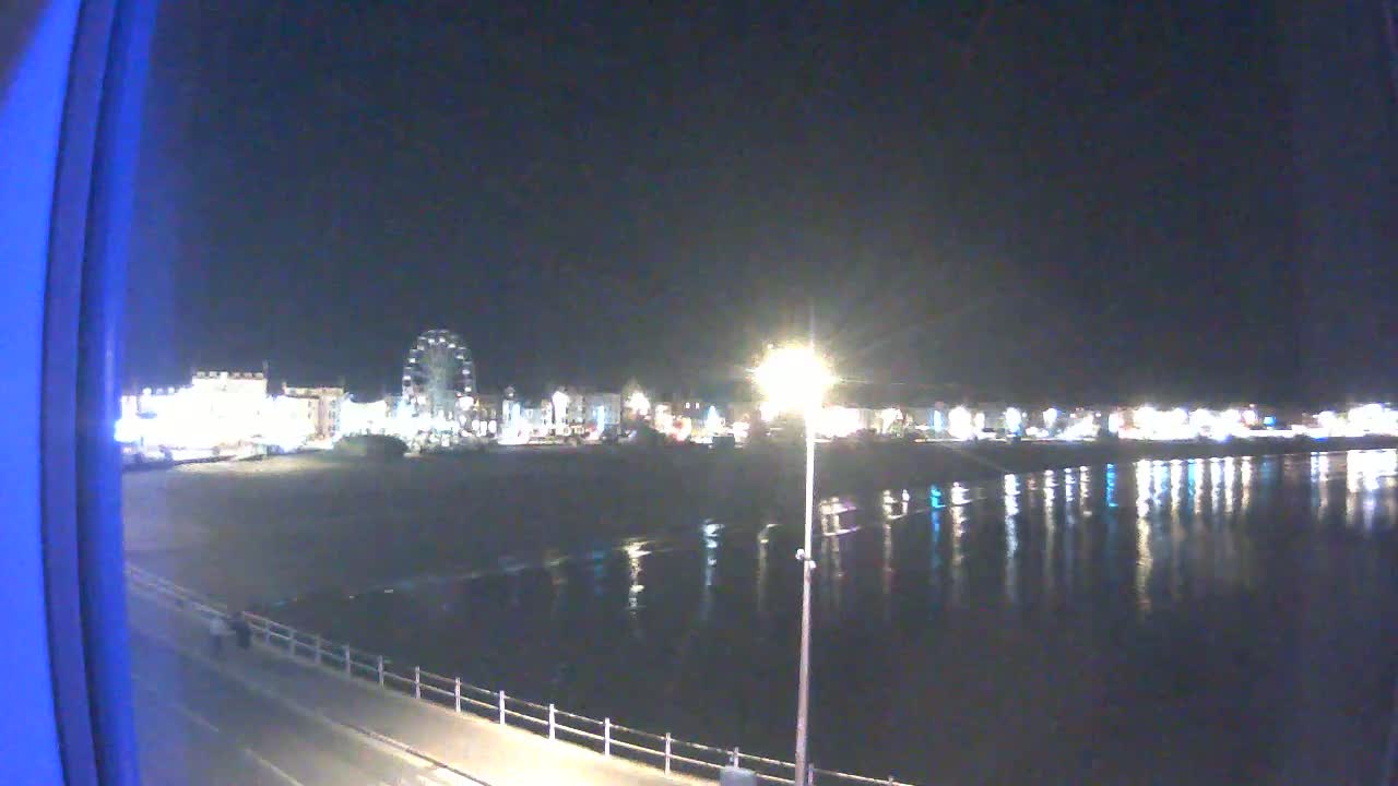 A nighttime view of a brightly lit seaside town with a Ferris wheel, seen across a calm, reflective body of water from a slightly elevated vantage point.