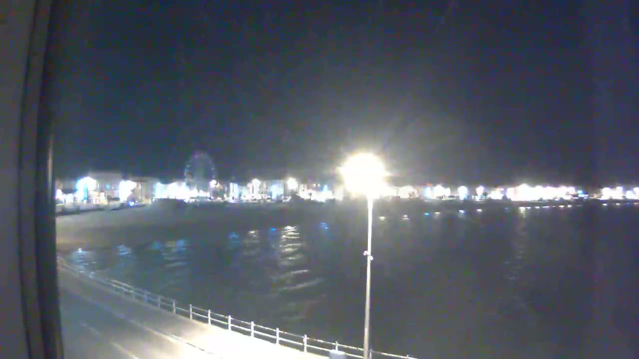 A nighttime view shows a dark body of water reflecting the lights of a town with a Ferris wheel, all under a clear, dark sky.