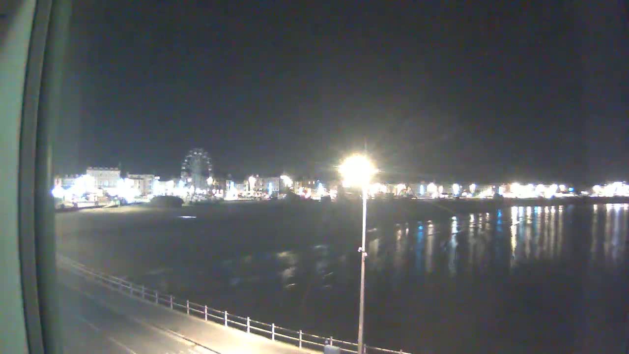 A nighttime view of a beach town's illuminated waterfront, featuring a Ferris wheel and calm water reflecting the lights.