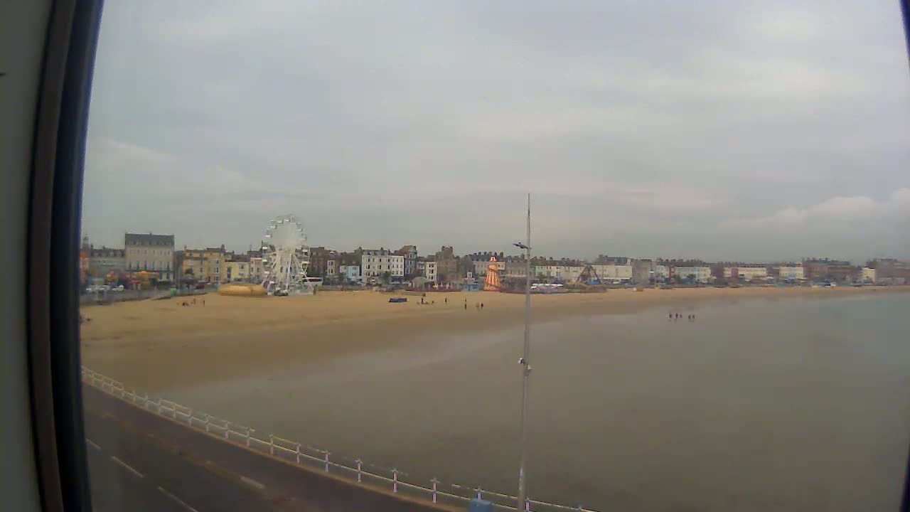 A Ferris wheel and several buildings are visible on a sandy beach next to calm, shallow water under an overcast sky.
