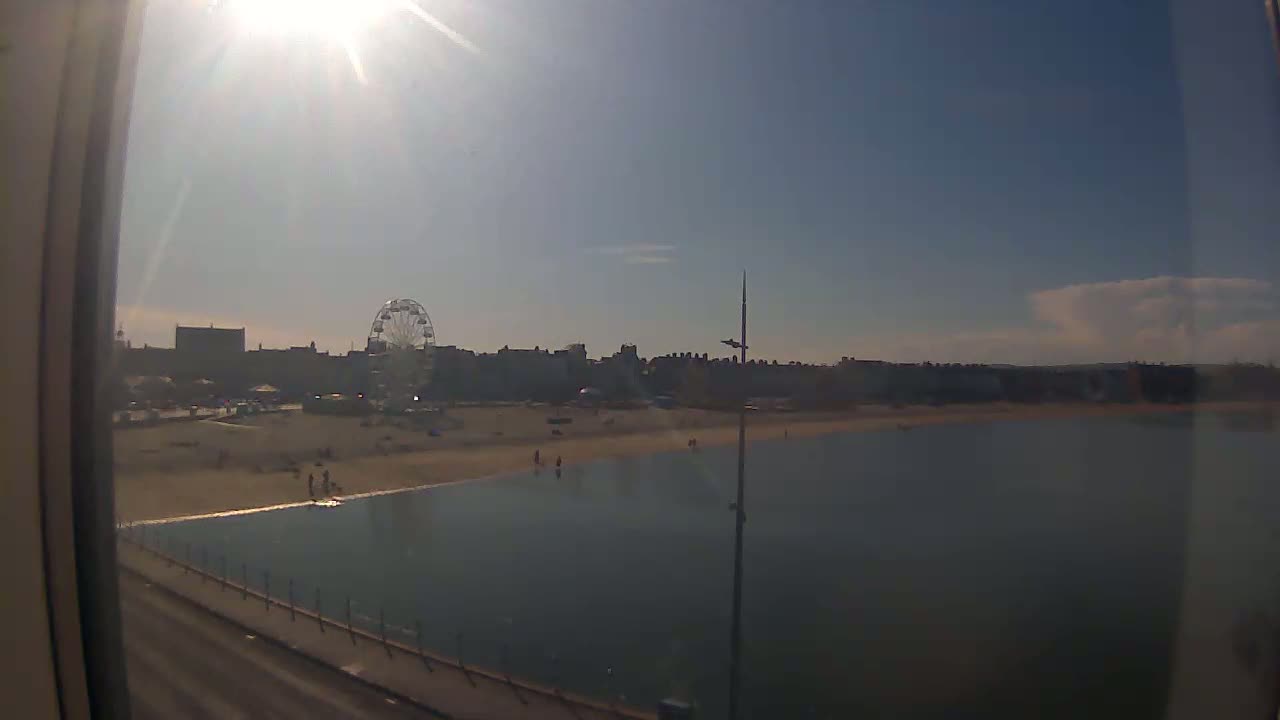 A sunny day at a beach shows a Ferris wheel and buildings in the distance, with calm water stretching towards the viewer.