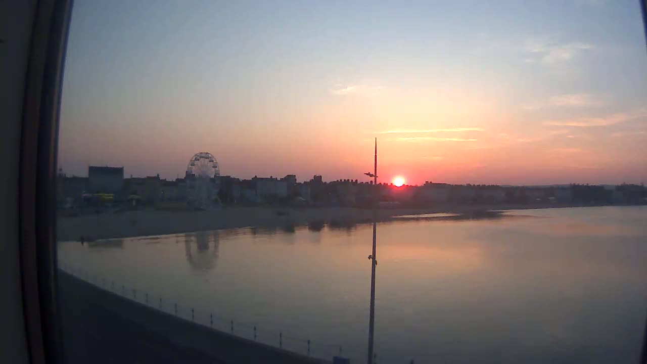 A calm body of water reflects a sunset over a beach town with a Ferris wheel.