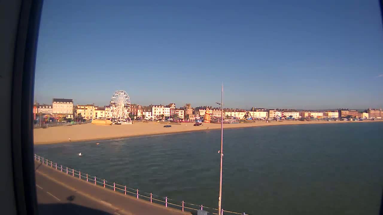 A sandy beach with a Ferris wheel and buildings behind it is visible from across calm water on a sunny day.