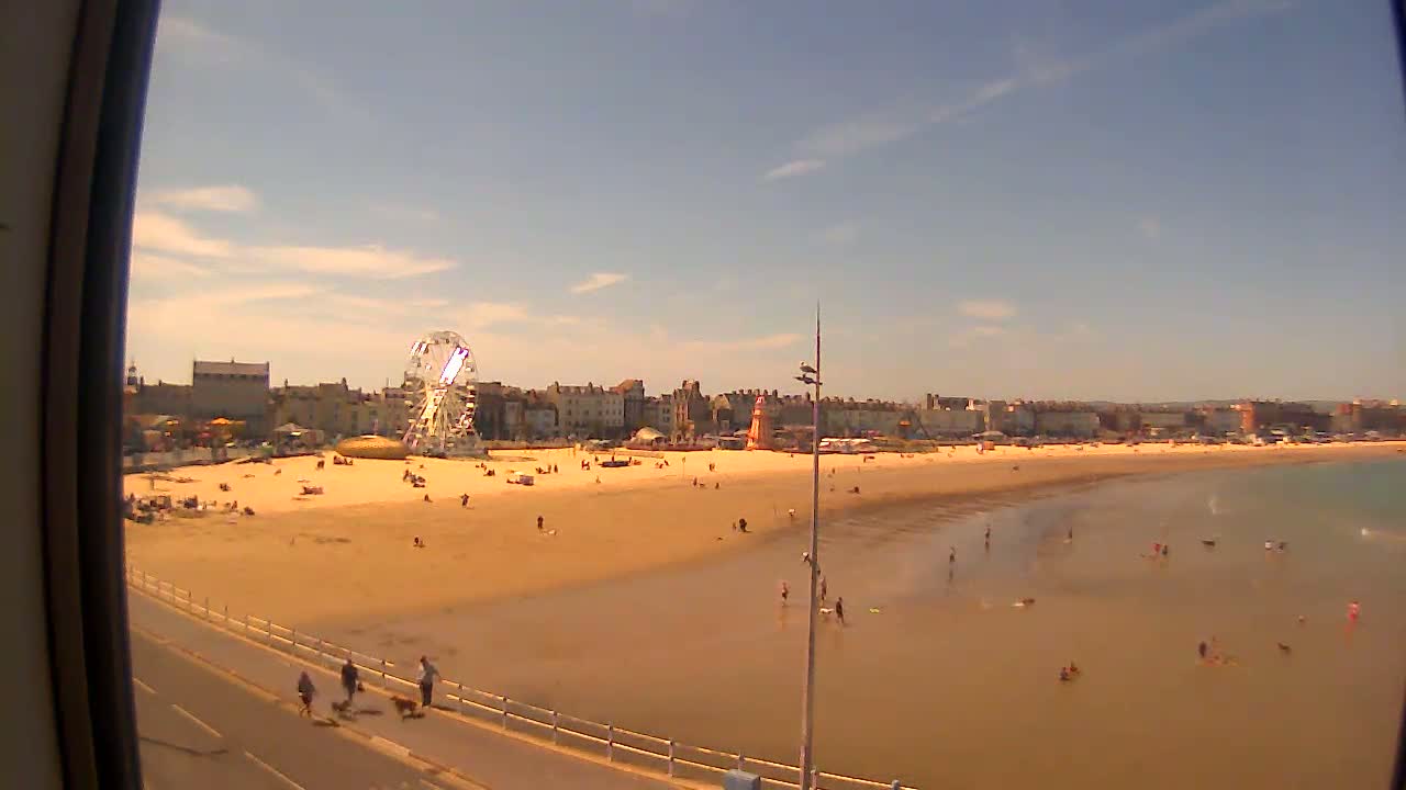 A sunny day at a beach with a Ferris wheel, people sunbathing and swimming in shallow water, and buildings along the shore.