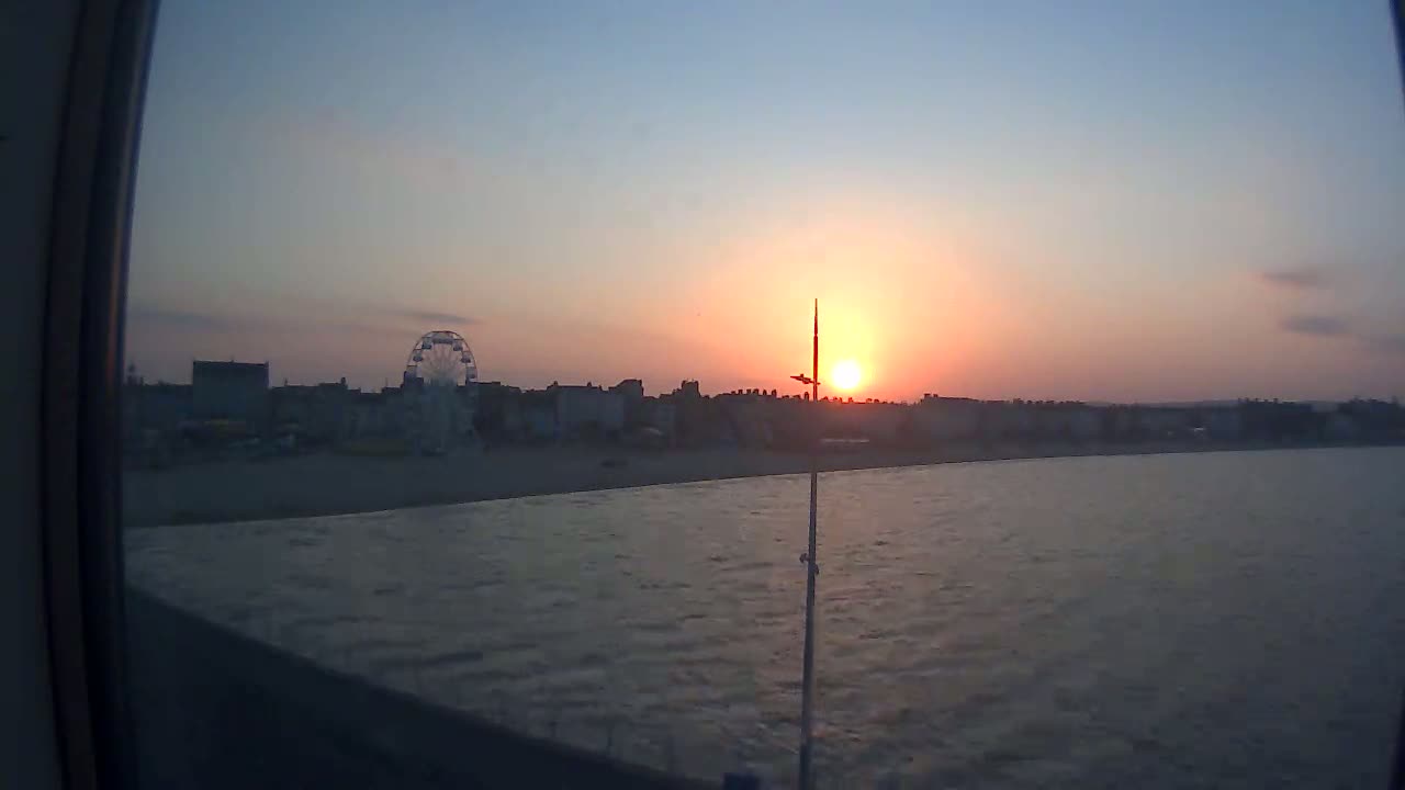 A seaside town with a Ferris wheel is silhouetted against a setting sun over calm water.
