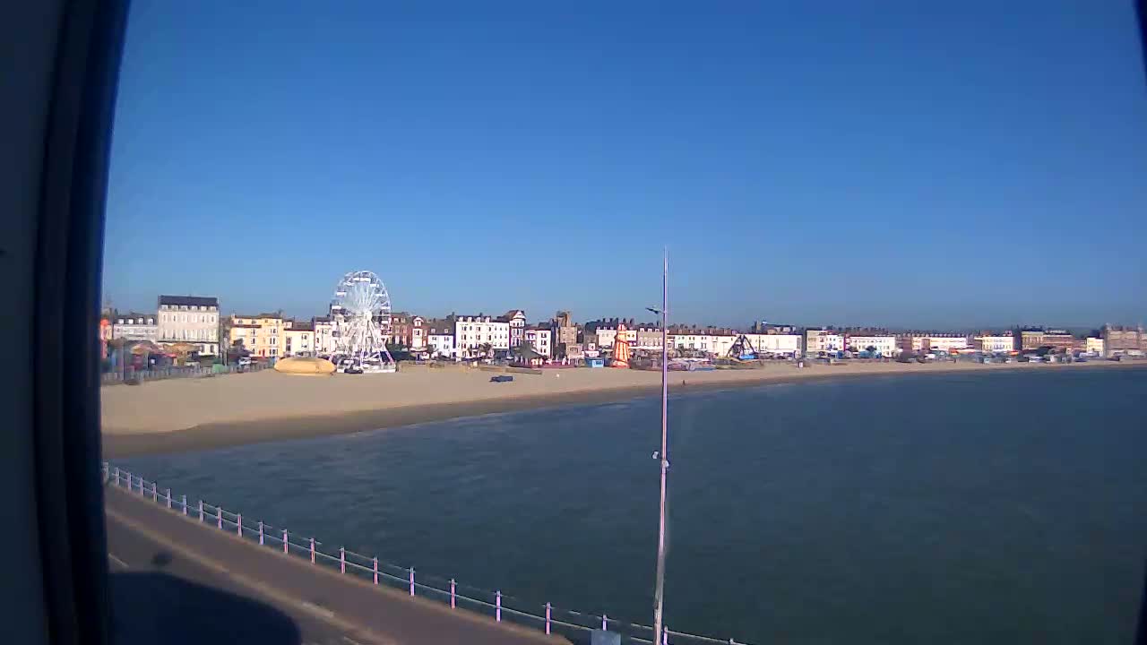 A Ferris wheel and beachfront buildings are visible on a sunny day, with calm water lapping a sandy shore.