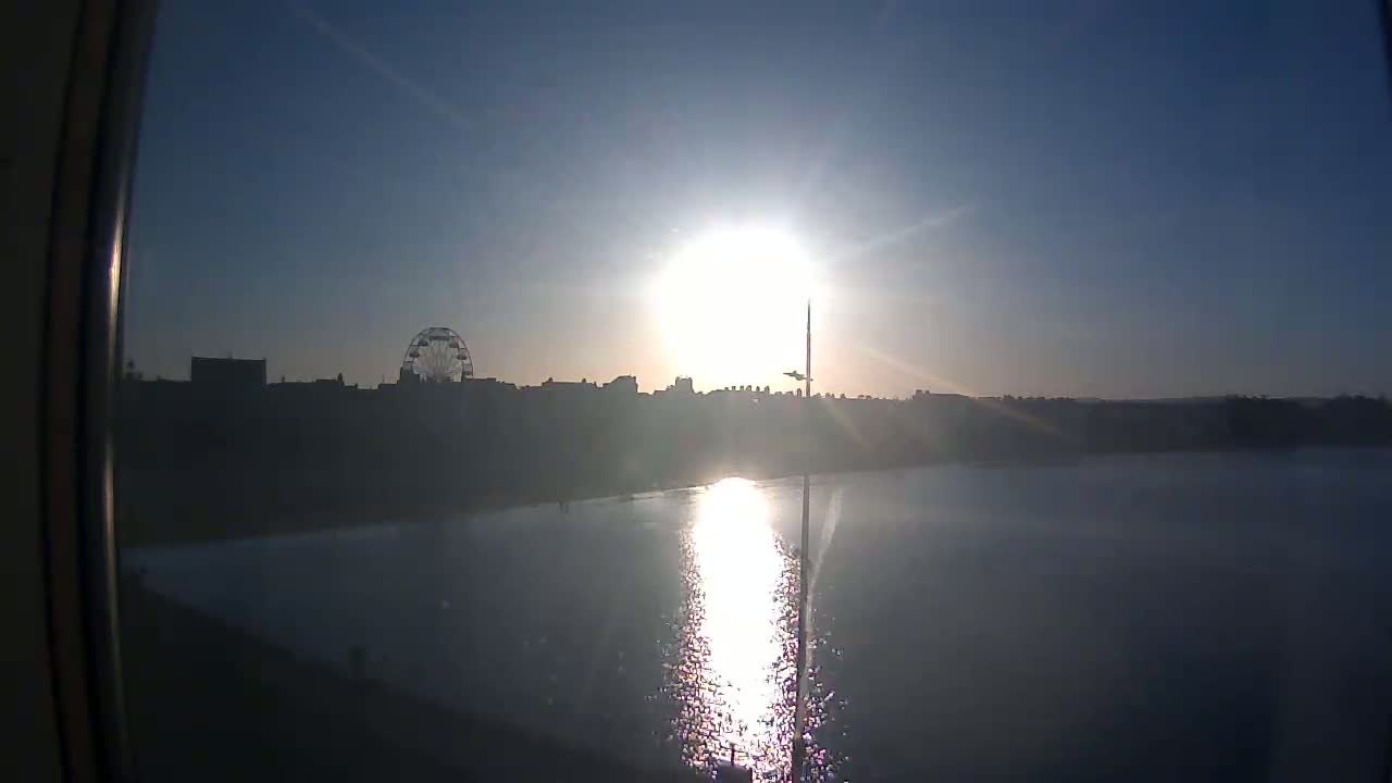 A bright sun shines over a calm body of water reflecting its light, with a distant city skyline silhouetted against the clear sky, featuring a Ferris wheel.