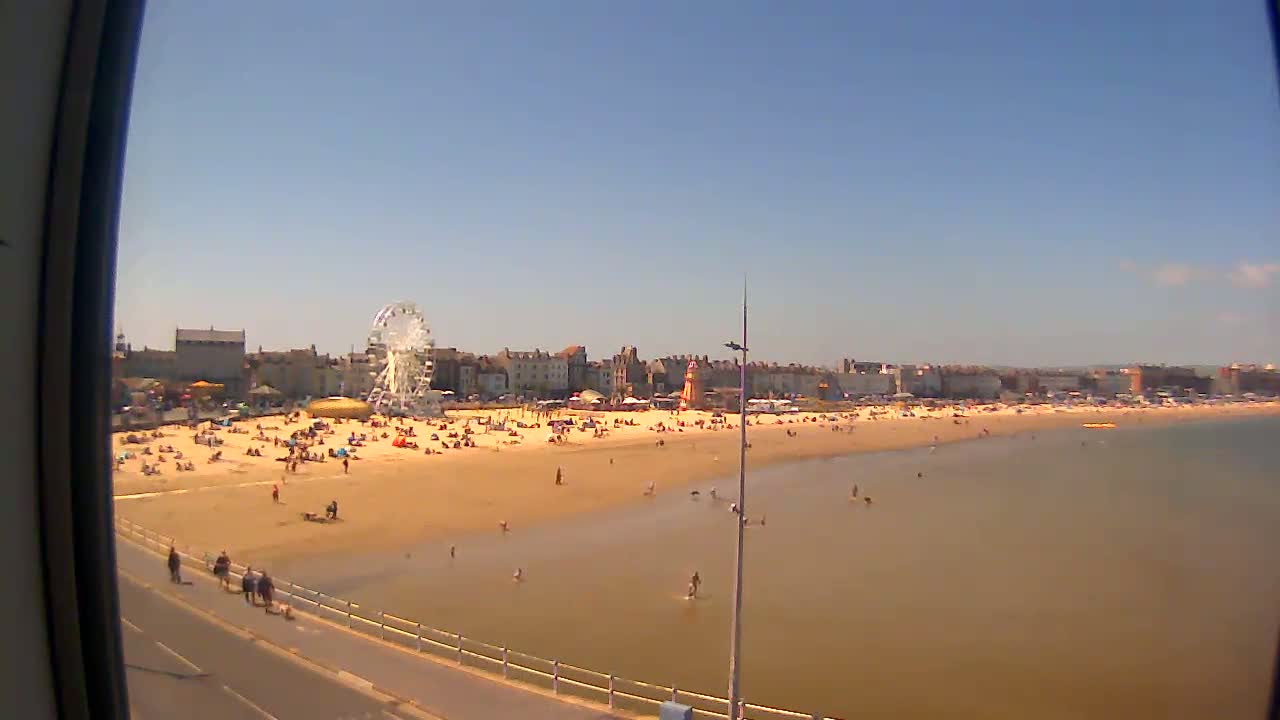 A sandy beach filled with people, a Ferris wheel, and buildings is visible on a sunny day with calm, shallow water.