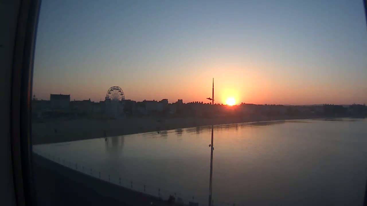 A beach town skyline silhouetted against a calm, orange sunset over a body of water, featuring a Ferris wheel.