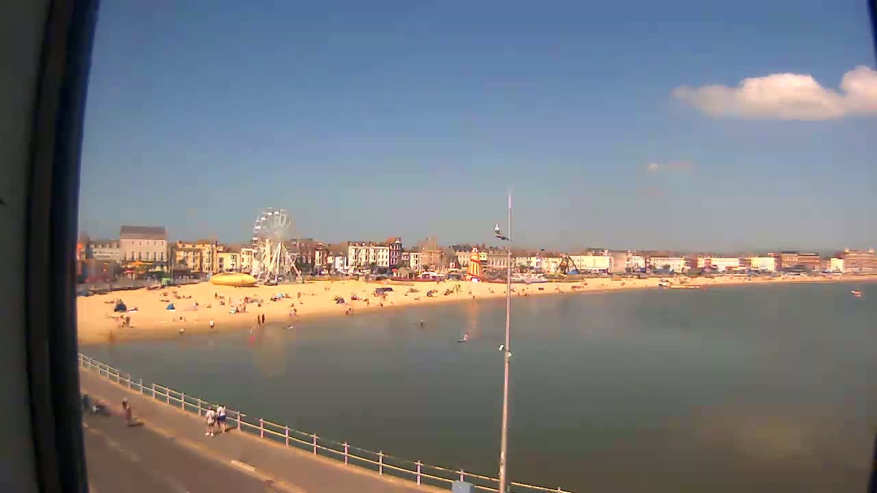 A sunny day shows a crowded beach with calm water, a Ferris wheel, and buildings along the shoreline.