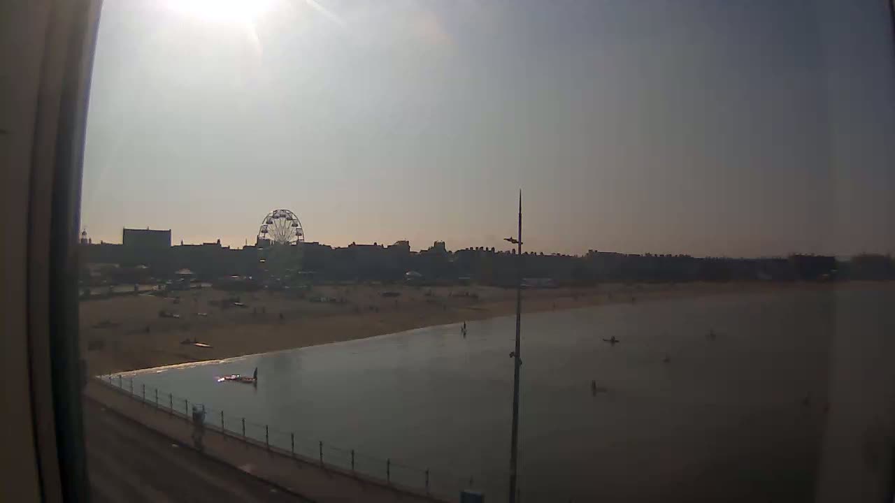 A sunny day at a beach shows a Ferris wheel and buildings in the background, with calm water in the foreground and a few people on the beach and in the water.