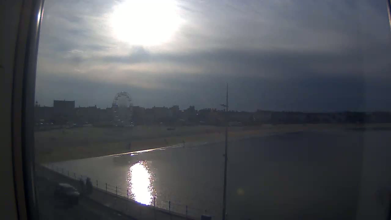 A partly sunny day shows a beach, calm water reflecting sunlight, and a Ferris wheel in the distance.