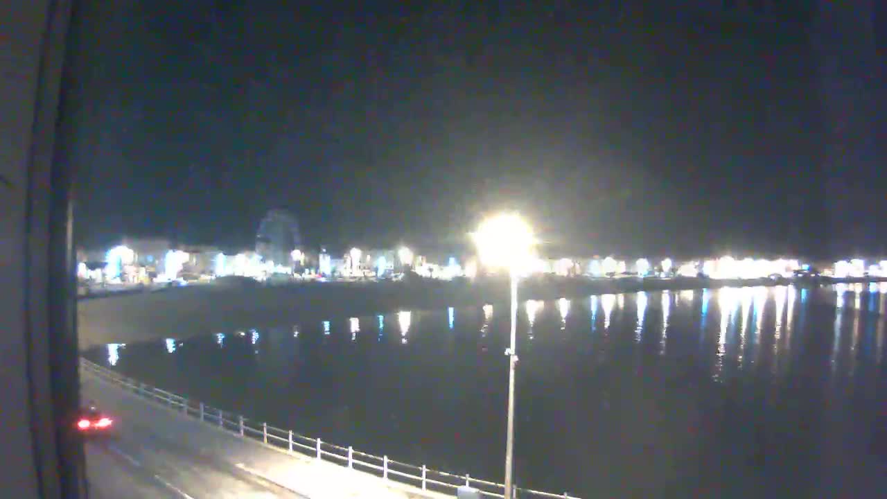 A nighttime view of a calm body of water reflecting the lights of a town across the water, with a road and a railing visible in the foreground.
