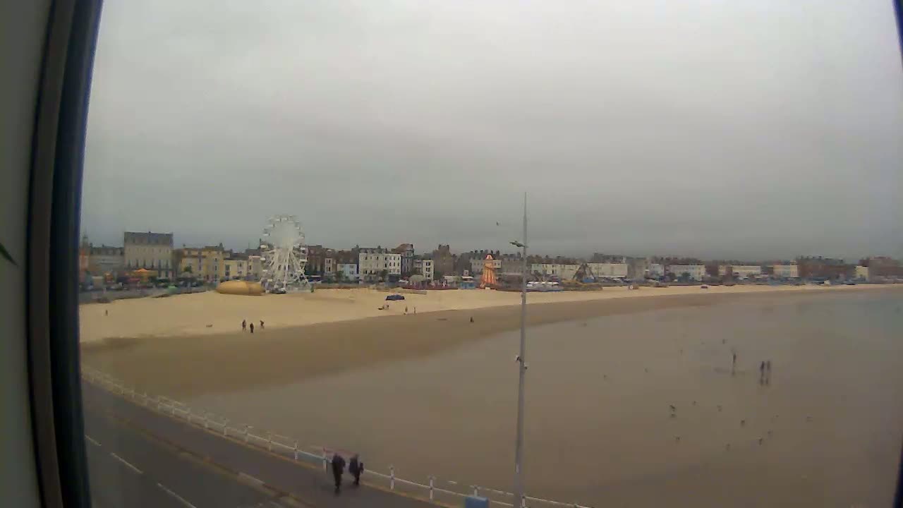 A wide shot shows a beach with a Ferris wheel and buildings in the background under an overcast sky.