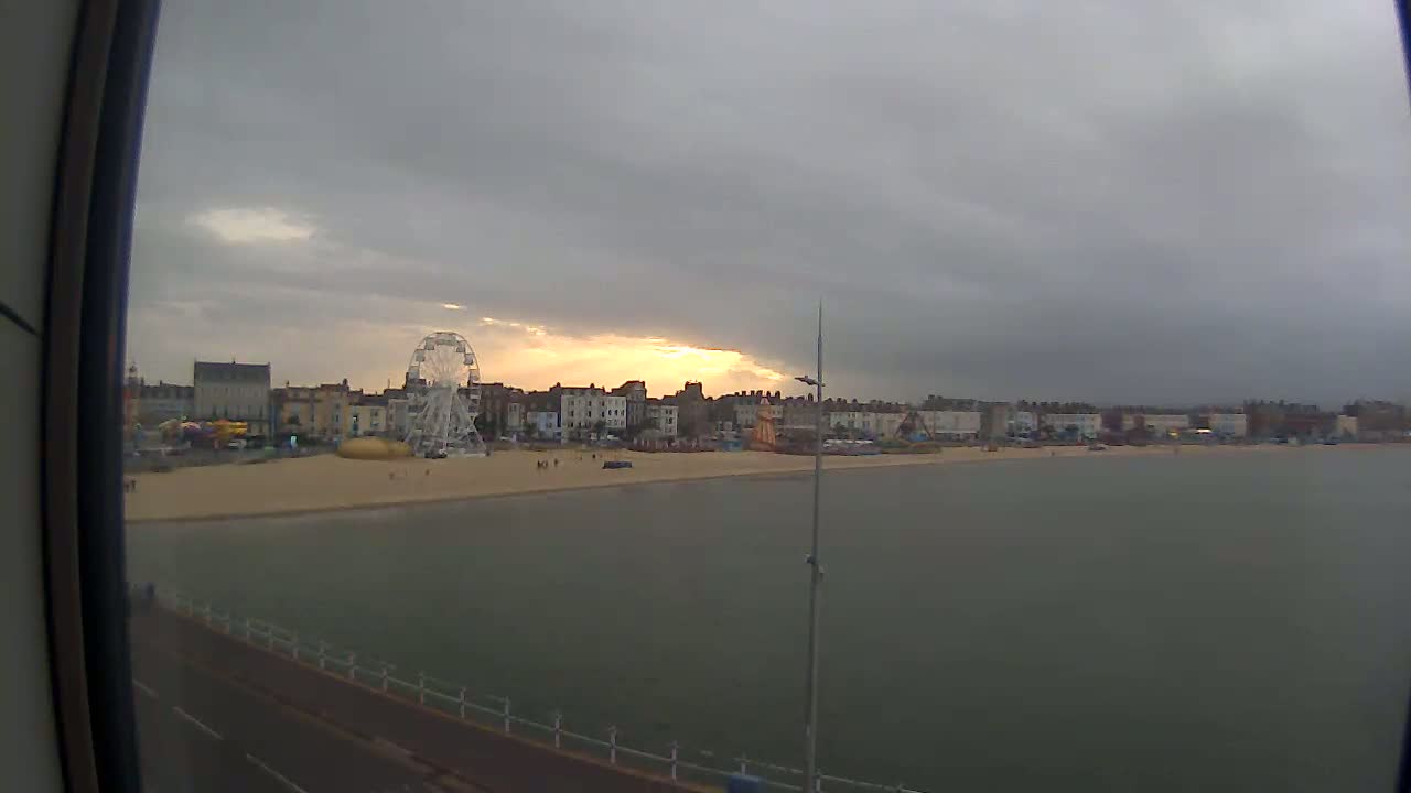 A beach with a Ferris wheel and buildings is seen under an overcast sky, with a calm sea in the foreground.