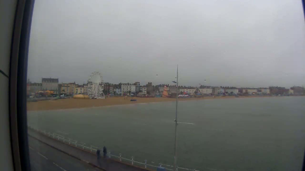 A seaside town with a Ferris wheel is visible across a calm, grey body of water under an overcast sky.