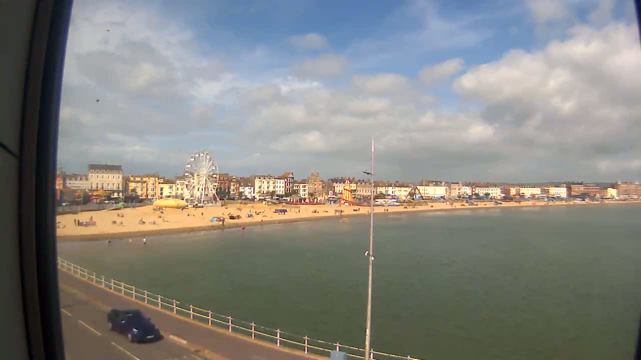 A partly cloudy day shows a beach with a Ferris wheel and other amusement park rides behind a row of buildings, and a calm body of water in front.