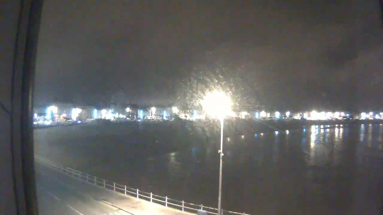 A nighttime view of a calm body of water reflecting the lights of a distant town under a slightly overcast sky.