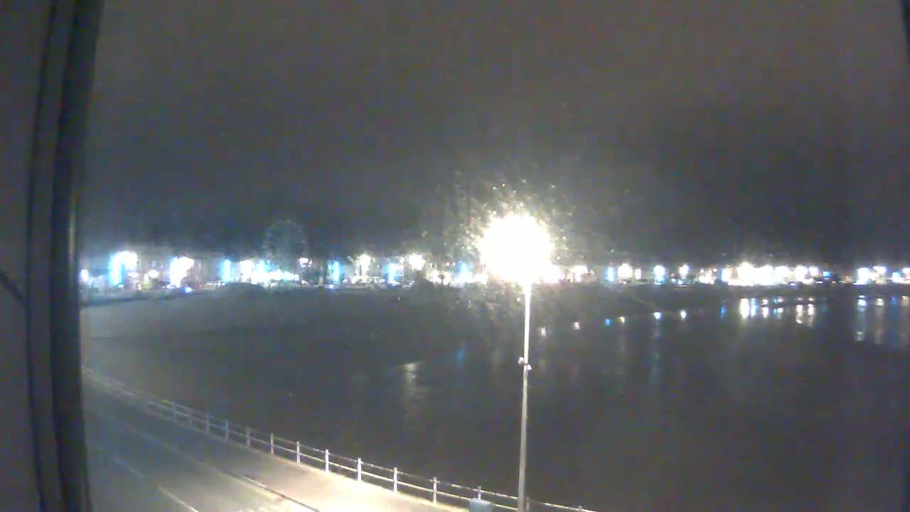 A nighttime view of a waterfront town, with lights reflecting on calm water under a dark, possibly overcast sky.