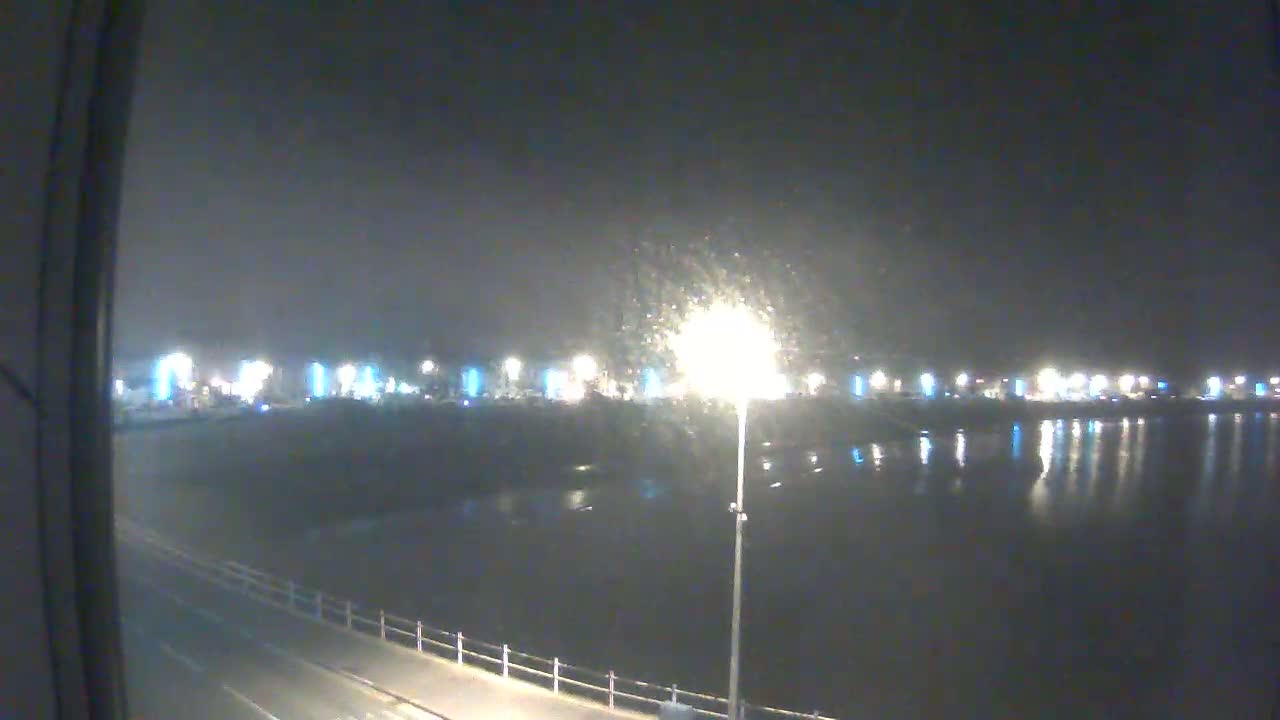 A nighttime view of a calm body of water reflecting the lights of a distant town under a slightly hazy sky.
