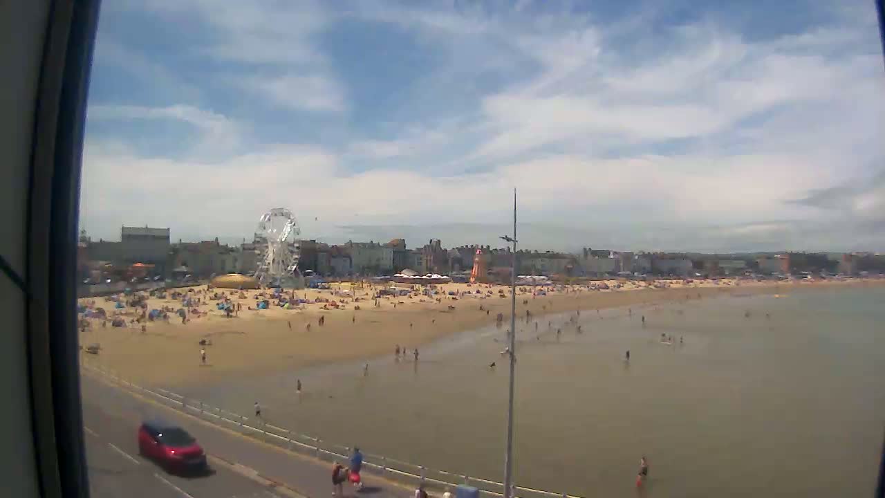A sunny day at a beach shows a sandy shore crowded with people, buildings, a Ferris wheel, and calm, shallow water.
