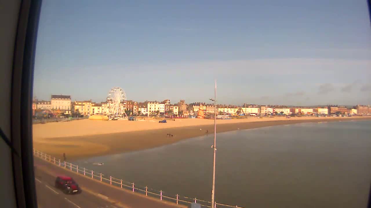 A sandy beach with a Ferris wheel and several buildings behind it stretches along calm, shallow water under a clear sky.