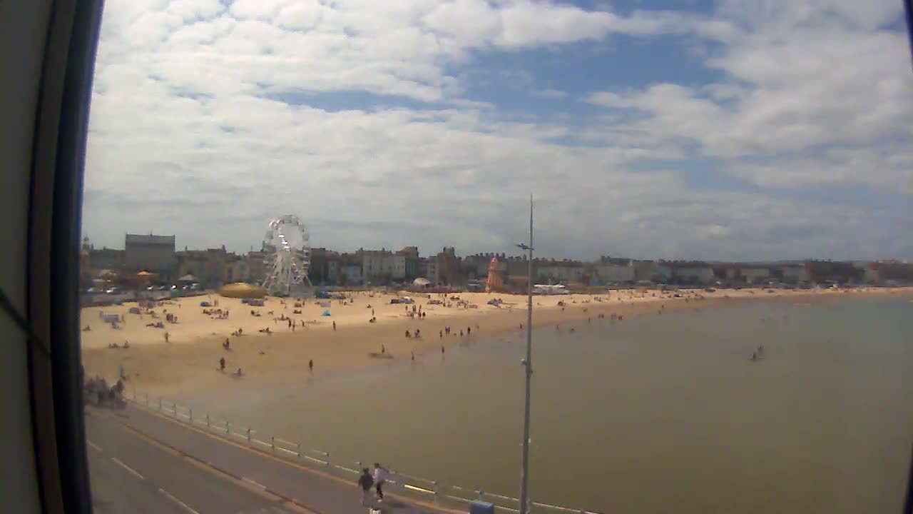 A partly cloudy day shows a beach scene with many people sunbathing and swimming in shallow water, a Ferris wheel and buildings in the background.