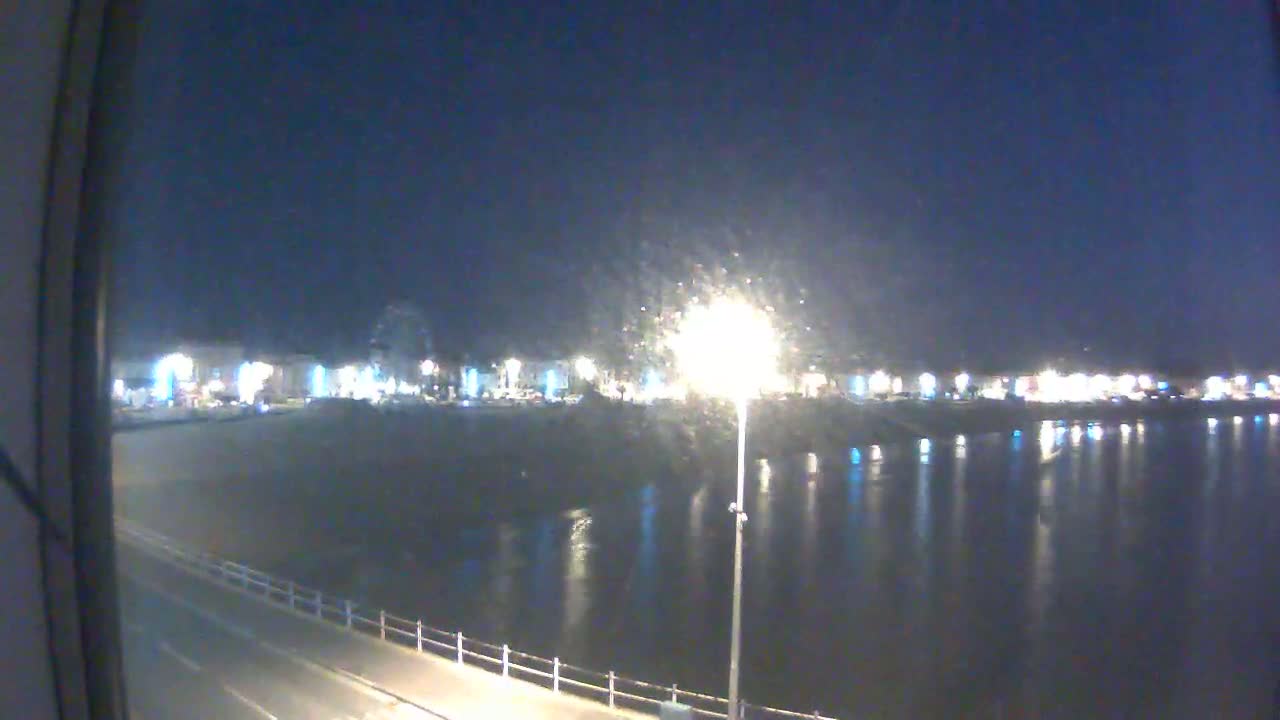 A nighttime view of a calm body of water reflecting the lights of a town across a paved waterfront.