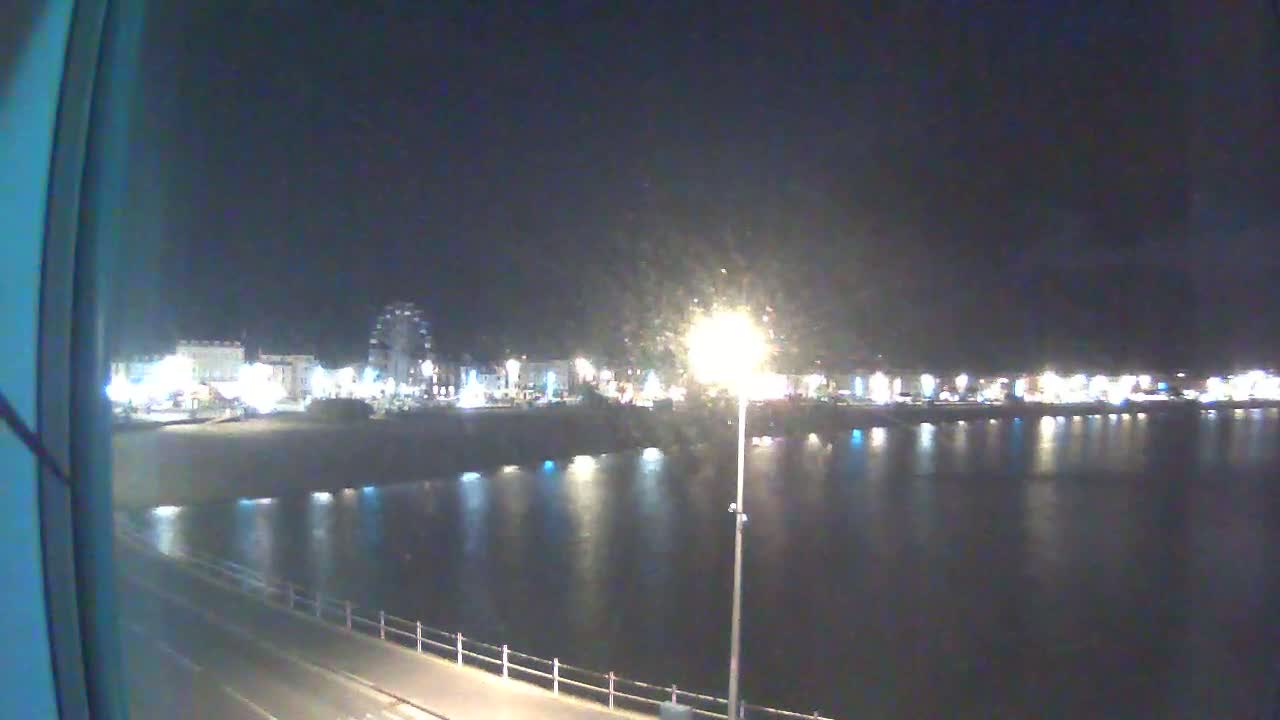 A nighttime view of a brightly lit waterfront town across a calm body of water, seen from a slightly elevated vantage point.
