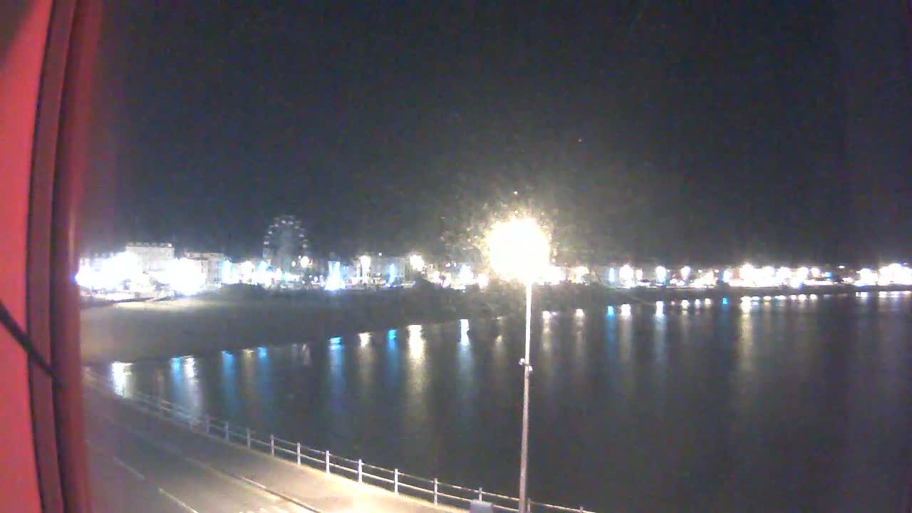 A nighttime view of a calm body of water reflecting the lights of a town across a beach and promenade.