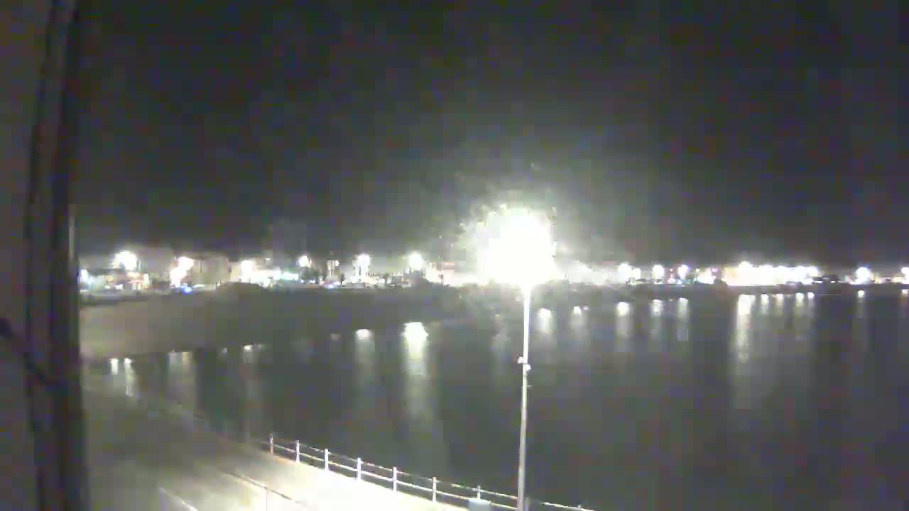 A nighttime view of a calm body of water reflecting the lights of a town across a bridge.