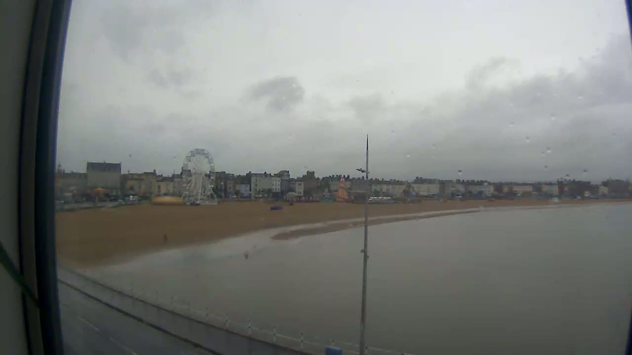 A rain-streaked view shows a Ferris wheel and buildings on a beach next to calm water.