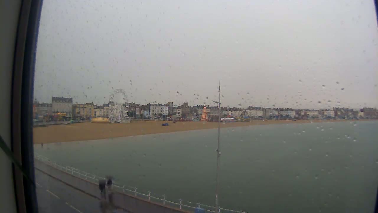 A rain-streaked view shows a beach with buildings and a Ferris wheel in the background, seen from what appears to be a window.