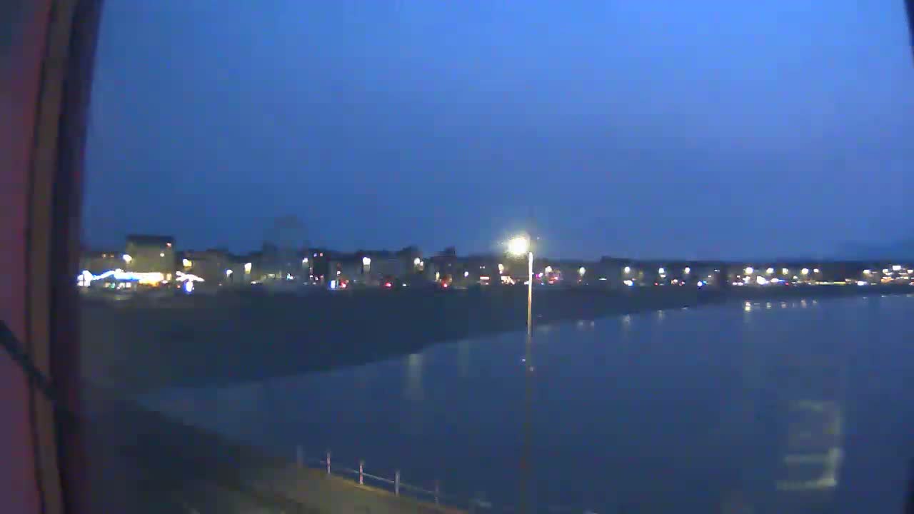 A nighttime view of a calm body of water reflecting the lights of a town across the shore under a dark blue sky.