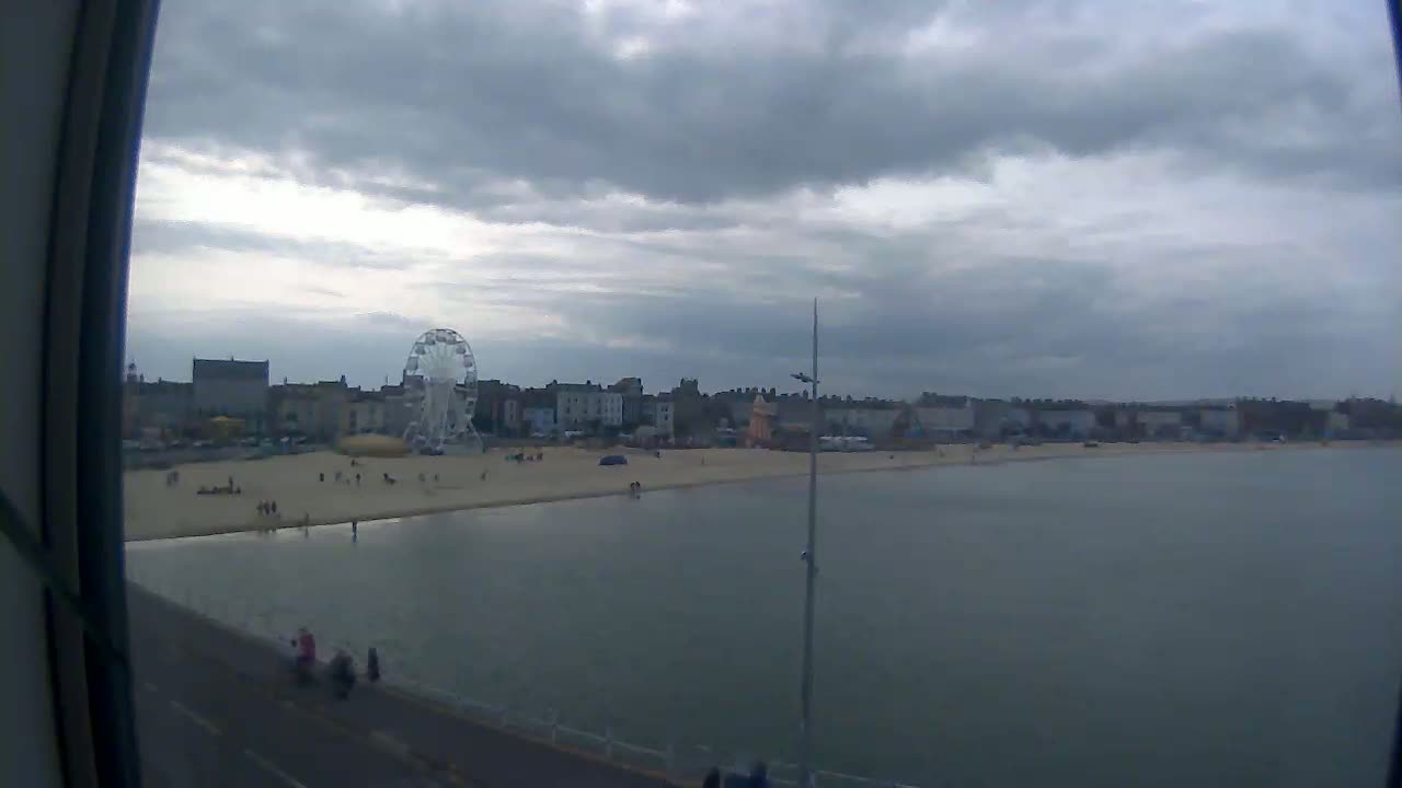 A seaside town with a Ferris wheel on a beach is viewed from across calm water under an overcast sky.