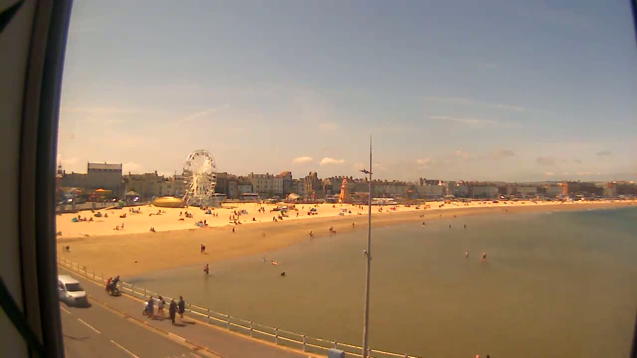 A sunny day at a beach shows a sandy shore with many people, a Ferris wheel, and buildings in the background.