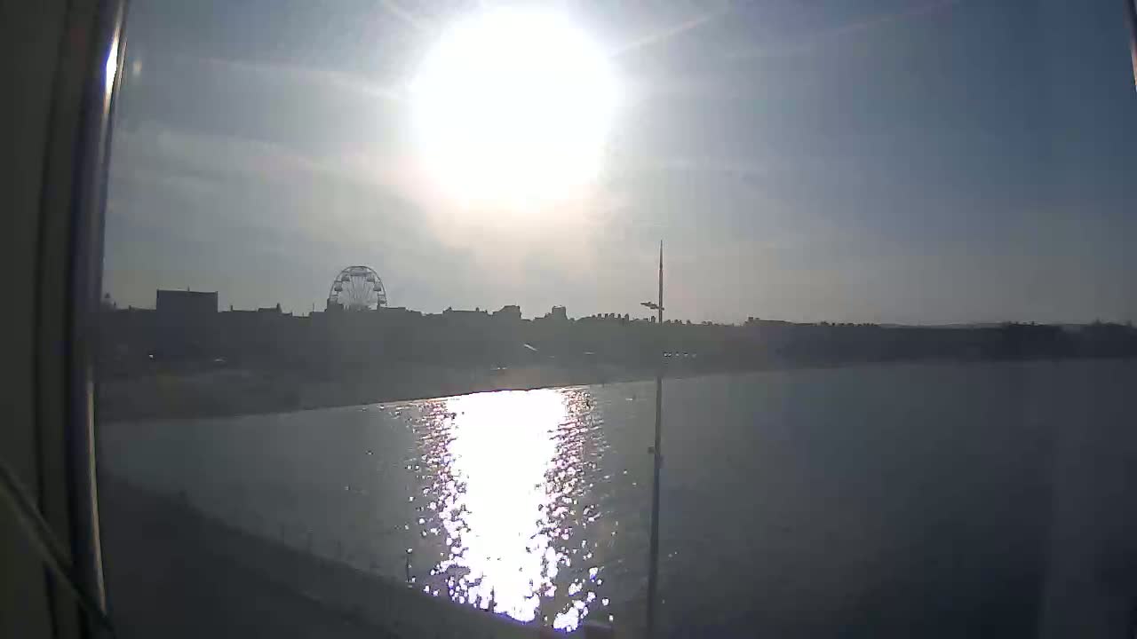 A bright, sunny day shows a body of calm water reflecting the sun, with a distant shoreline featuring a Ferris wheel and buildings.