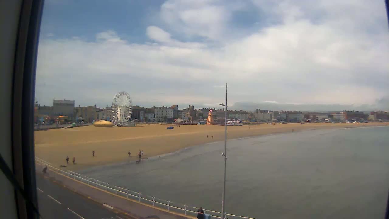 A sandy beach with a Ferris wheel and other amusement park rides, under a partly cloudy sky.